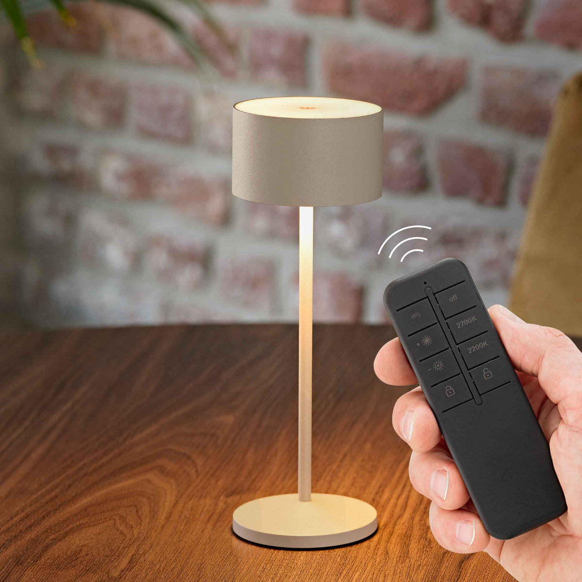 Sigor Veluna Remote Akku-Tischleuchte in Dünenbeige auf einem Holztisch, daneben eine Hand mit der Funkfernbedienung.