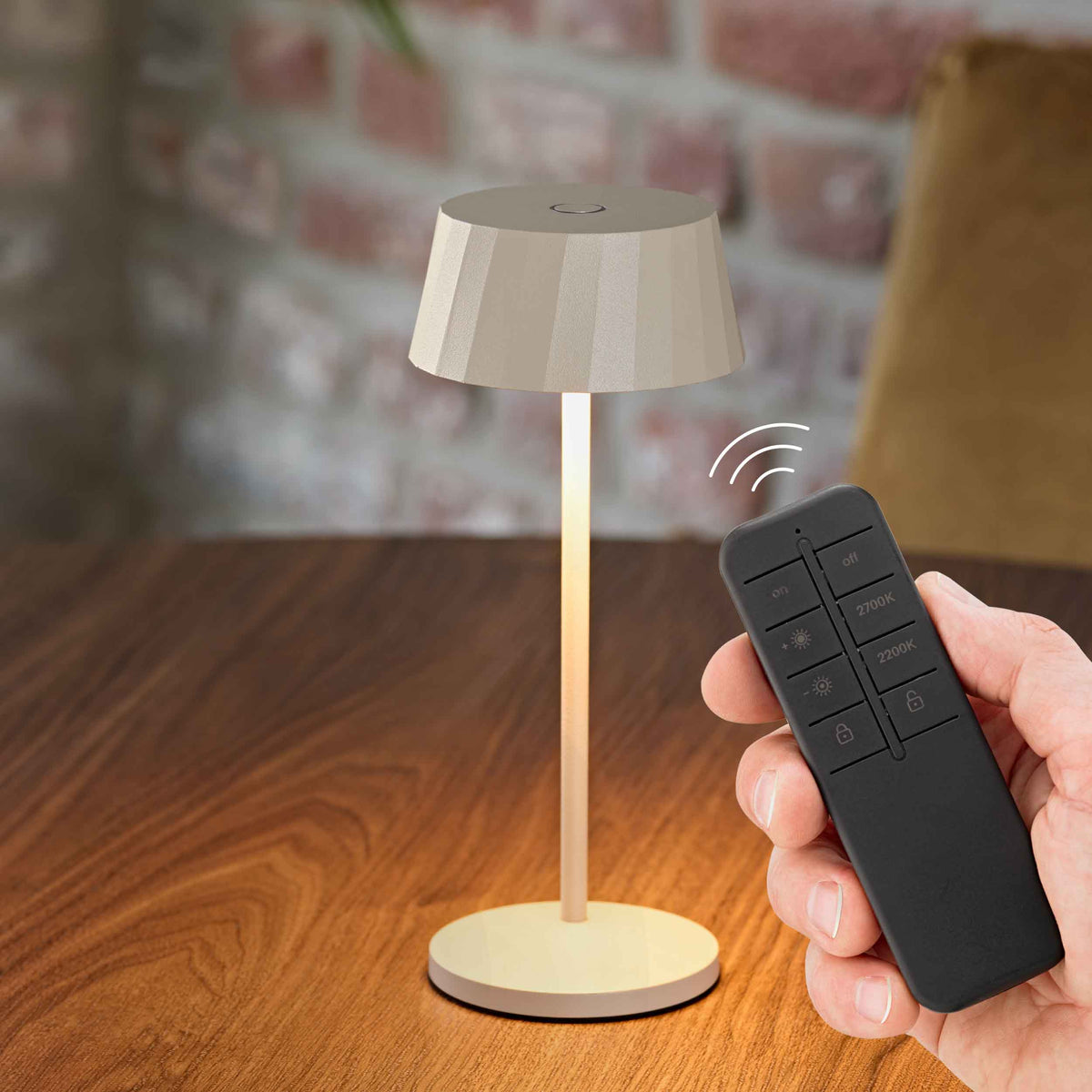 Sigor Facetta Remote Akku-Tischleuchte in Dünenbeige auf einem Holztisch, daneben eine Hand mit der Funkfernbedienung.